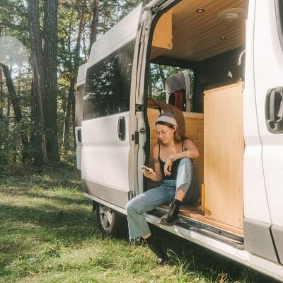Woman sitting in camper van in forest and using smartphone
